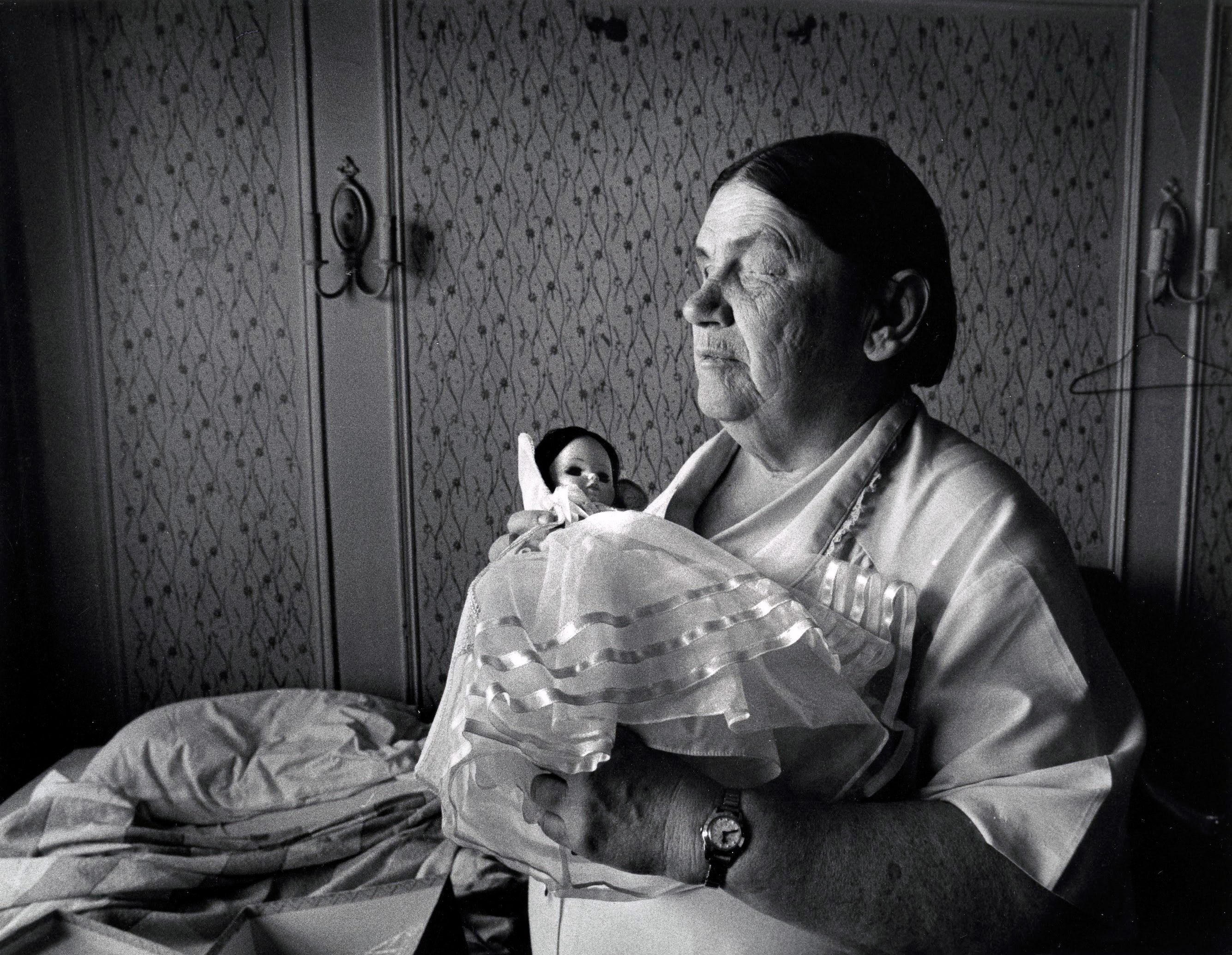 Elderly woman holding a doll, looking contemplative.